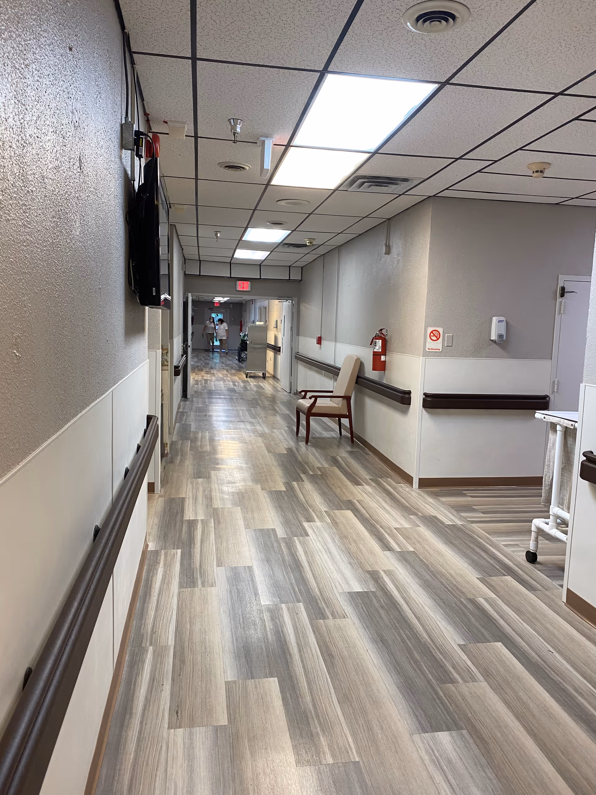 A long, well-lit hallway in a nursing and rehabilitation facility with wood-patterned flooring, beige walls, handrails on both sides, a chair against the right wall, a fire extinguisher, and a person in the distance near the end of the corridor.
