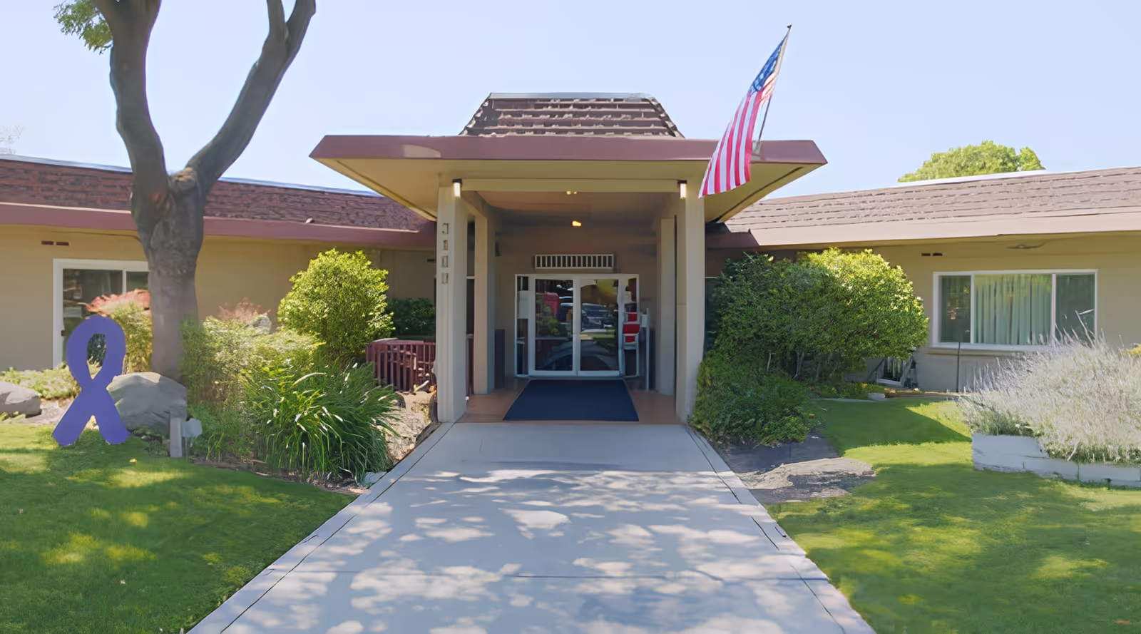 Entrance to a single-story care facility with a covered walkway, American flag, and landscaped lawn.
