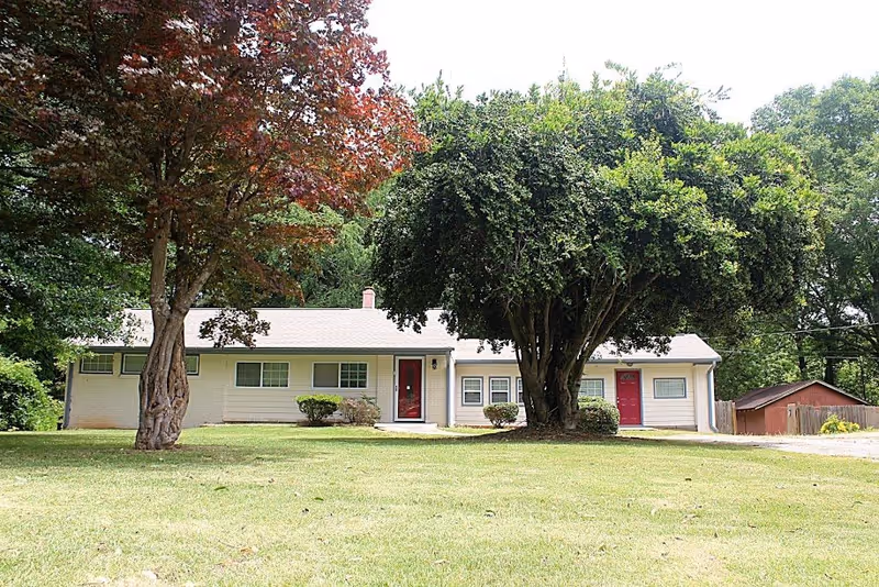 Single-story cream-colored house with two red doors, a large front lawn and mature trees.