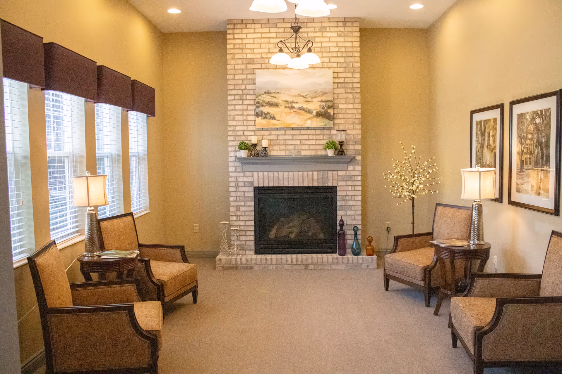 A cozy living room in a senior facility with a brick fireplace, seating, and artwork.