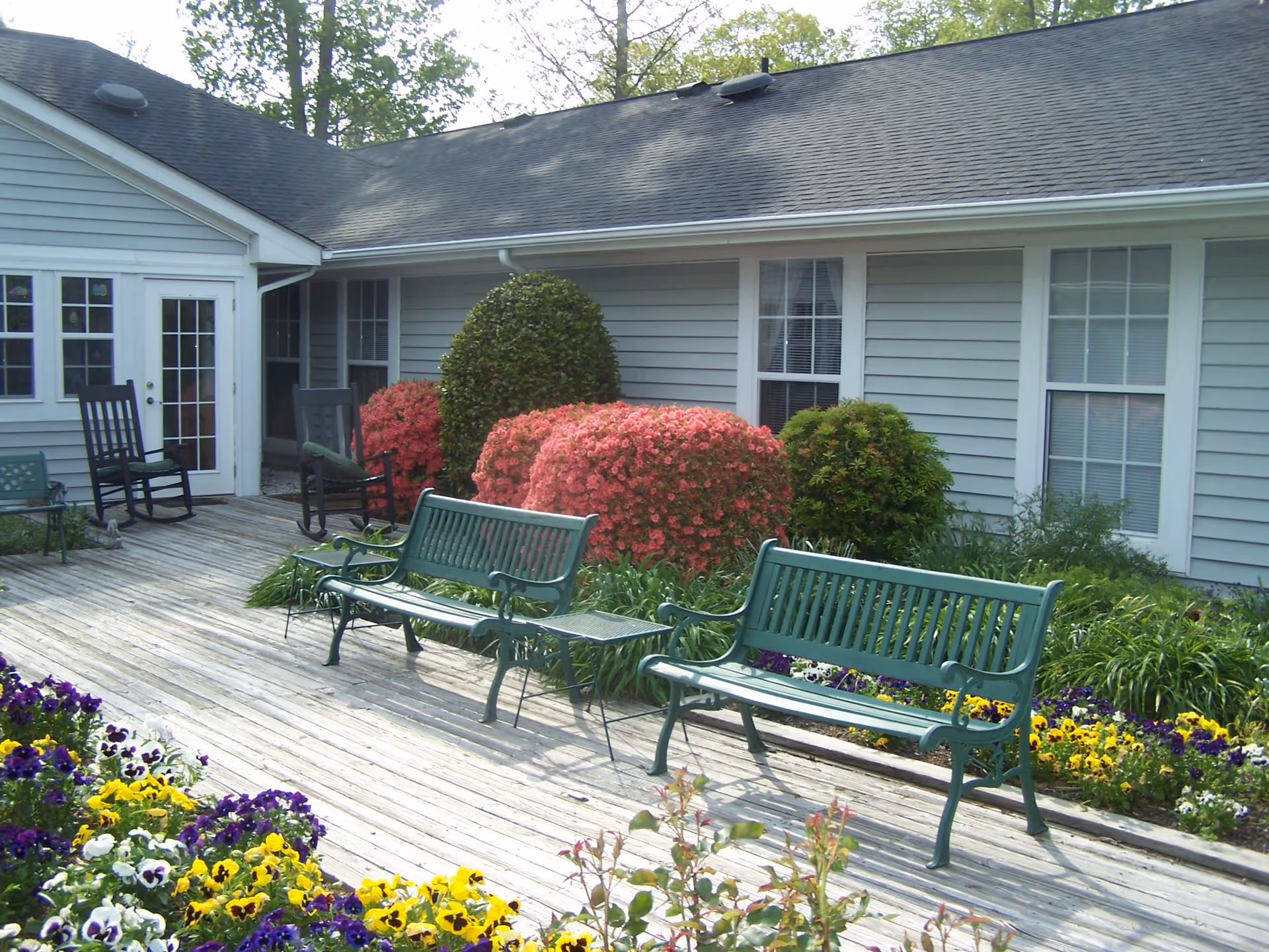 Wooden courtyard with green benches, rocking chairs, and colorful flowerbeds in front of a light-gray single-story building.