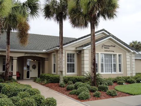 Front exterior of Summerfield Suites Assisted Living with palm trees and a landscaped entry.