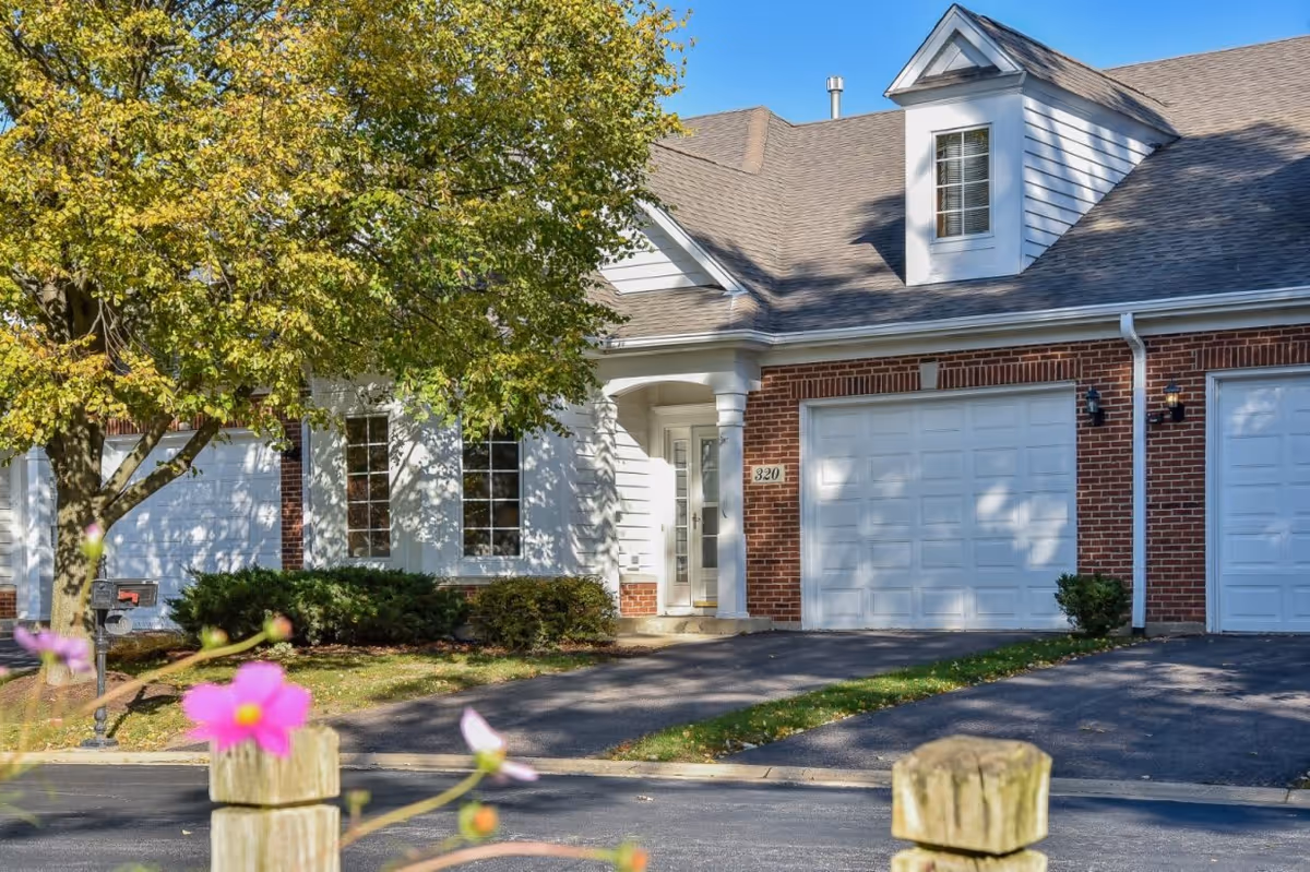 Exterior view of a residential-style building with a brick facade and white garage doors, surrounded by greenery including a tree with green leaves and some pink flowers in the foreground. The building has a pitched roof with dormer windows and a driveway leading to the garage.