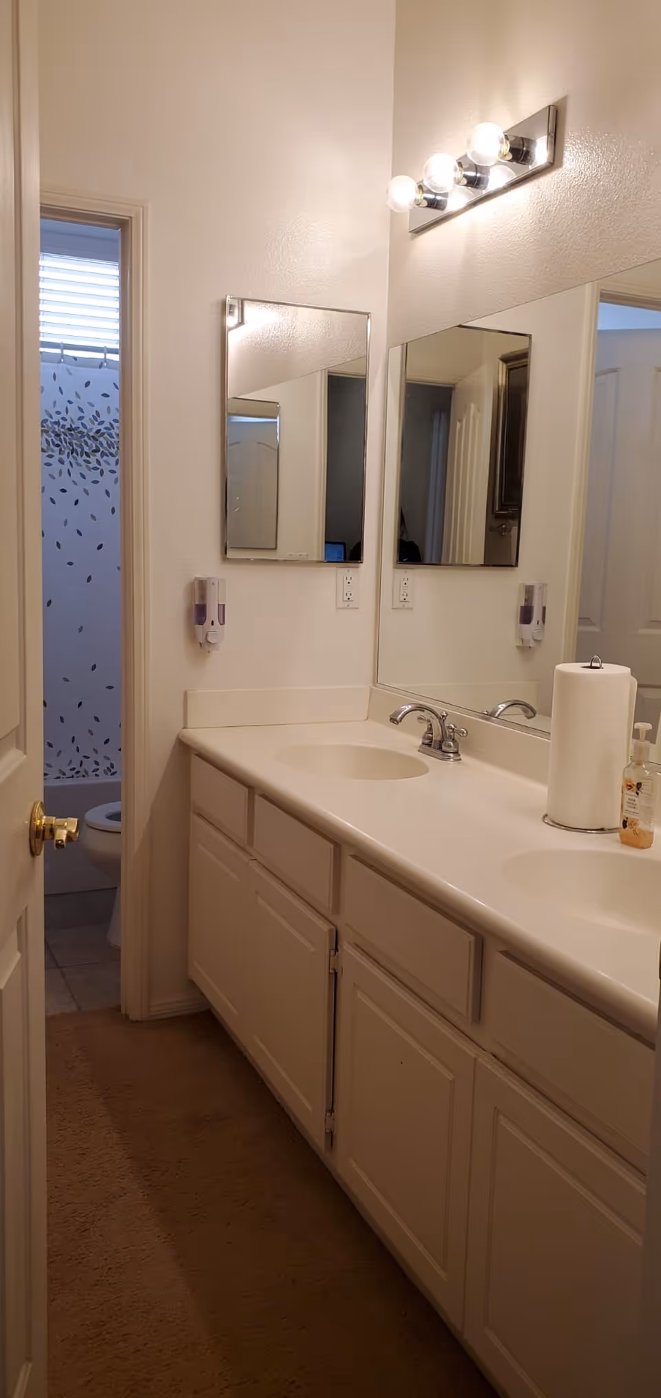 A bathroom with a double sink vanity featuring white countertops and cabinets. Above the sinks are two mirrors and a light fixture with four bulbs. To the left, there is a doorway leading to a toilet and a shower with a curtain decorated with a leaf pattern. A paper towel roll and a soap dispenser are on the countertop.