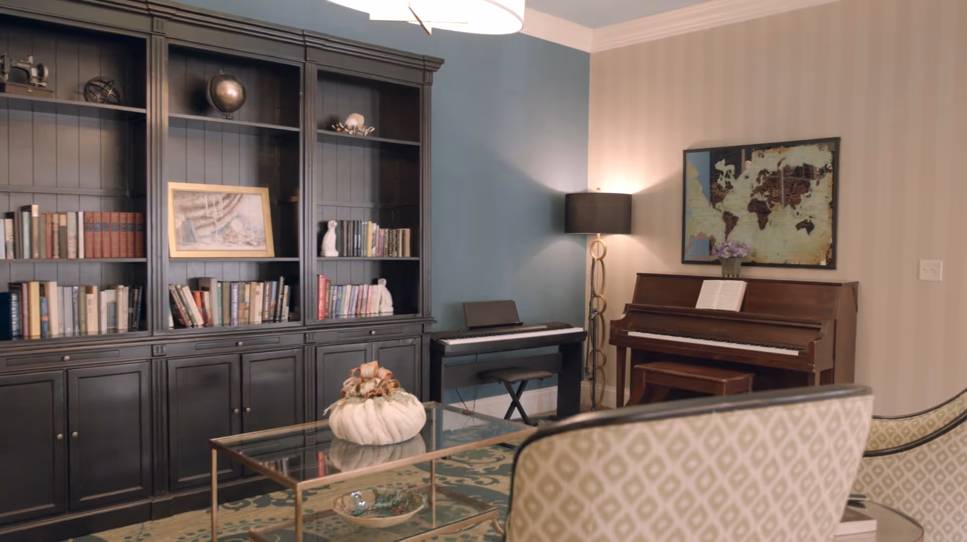 A cozy living room area with a large black bookshelf filled with books and decorative items, a glass coffee table with a white decorative pumpkin, a digital piano with a stool, an upright wooden piano with sheet music, a floor lamp with a black shade, and a framed world map on the wall.