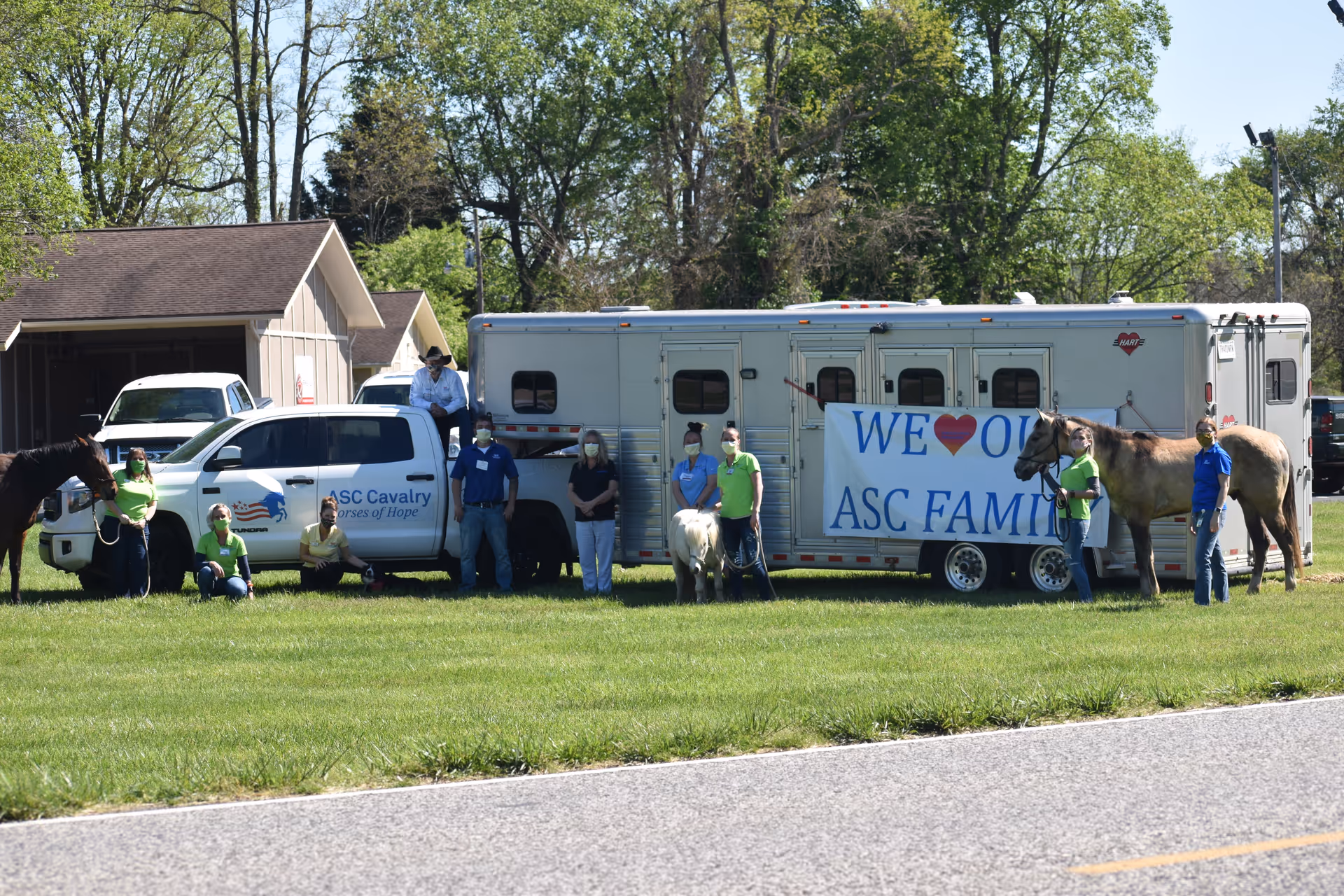 A group of people standing and sitting on grass in front of a white truck and horse trailer. The truck has a logo that reads 'ASC Cavalry Horses of Hope.' There are three horses present, and a large banner on the trailer reads 'WE ♥ OUR ASC FAMILY.' The scene is outdoors with trees and buildings in the background.