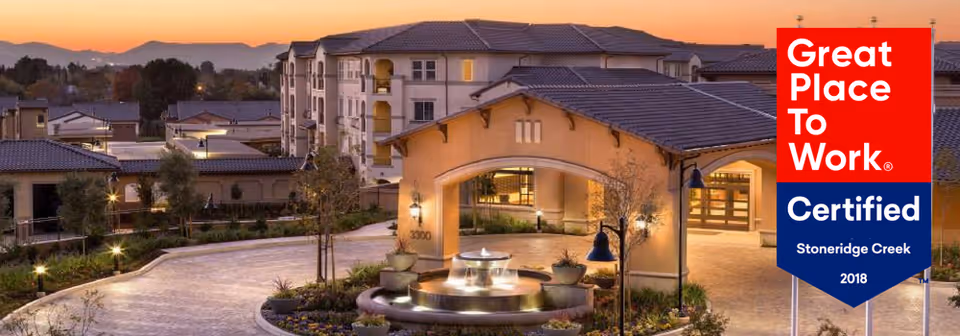 Front exterior of Stoneridge Creek Pleasanton at dusk showing the entrance, a circular fountain, and surrounding buildings.