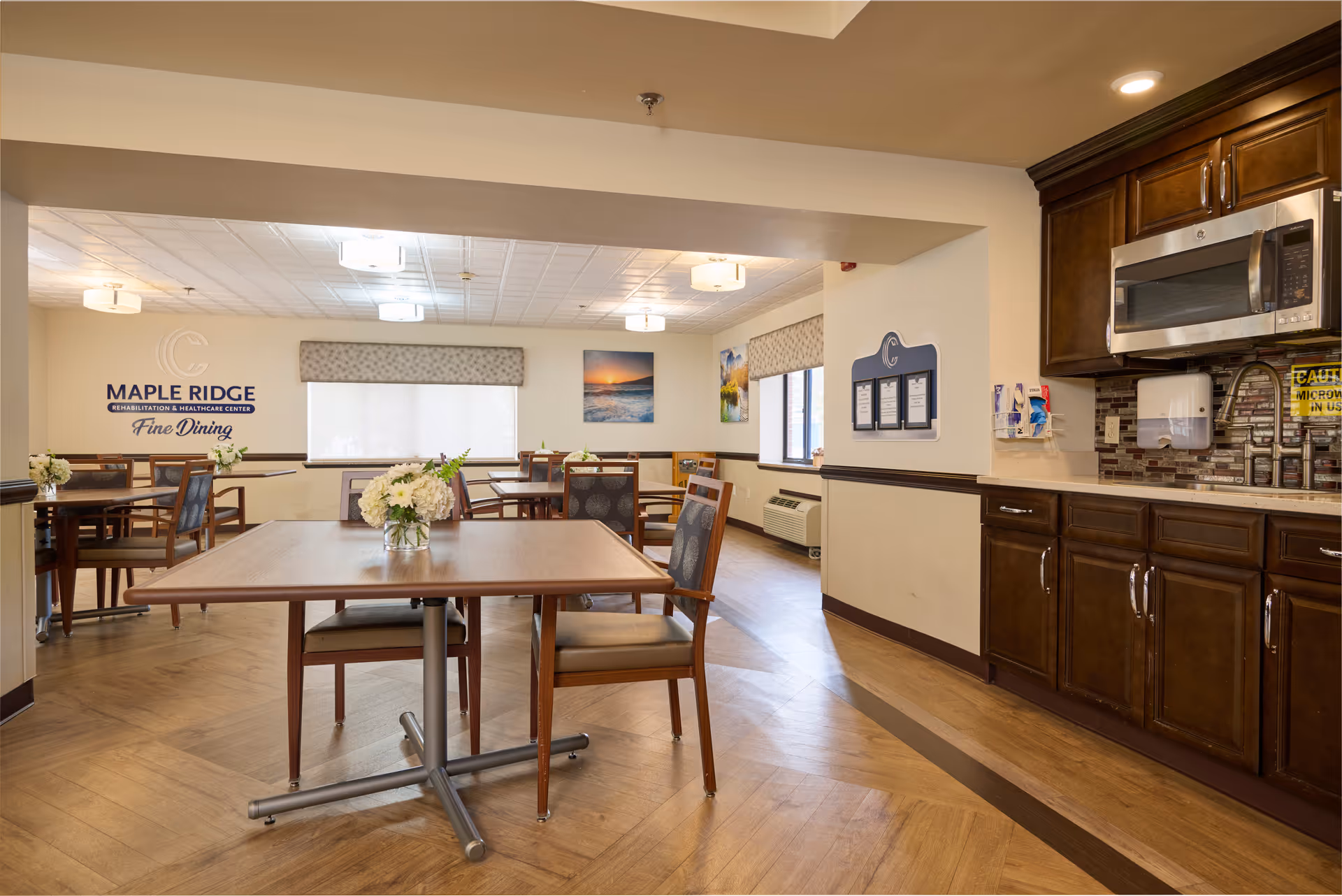 A dining area in Maple Ridge Rehabilitation and Healthcare Center featuring wooden tables and chairs with floral centerpieces. The room has large windows with blinds, wall art, and a kitchen area with dark wood cabinets, a microwave, and a sink. The wall displays the facility's name and the words 'Fine Dining'.
