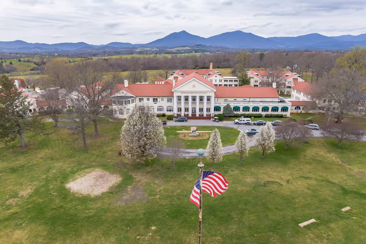 Aerial view of a large historic building with white columns and a red roof, surrounded by green lawns and trees with white blossoms. An American flag is prominently displayed in the foreground, and mountains are visible in the background under a cloudy sky.
