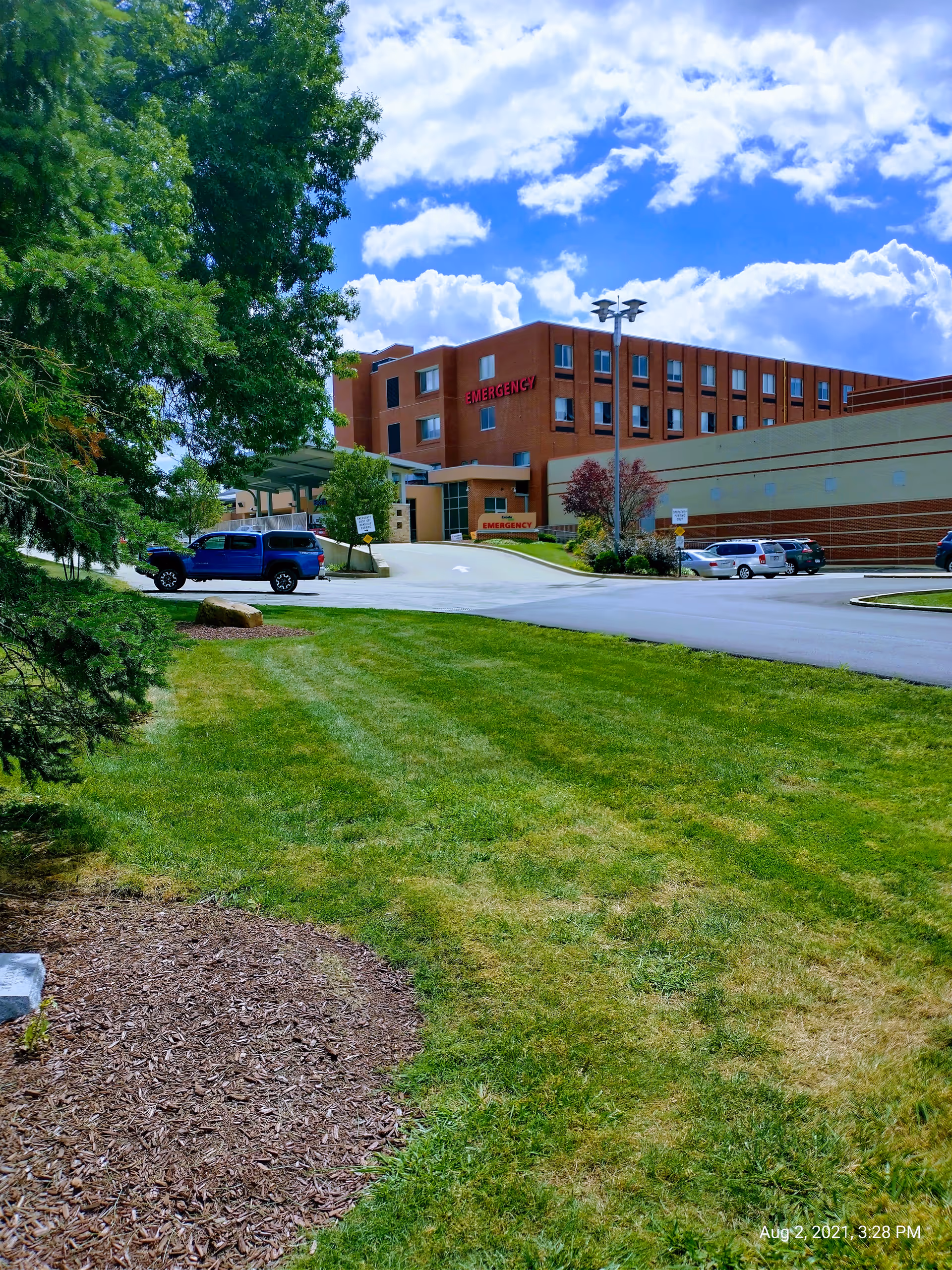 Brick hospital building with emergency entrance, parking lot, and green lawn.