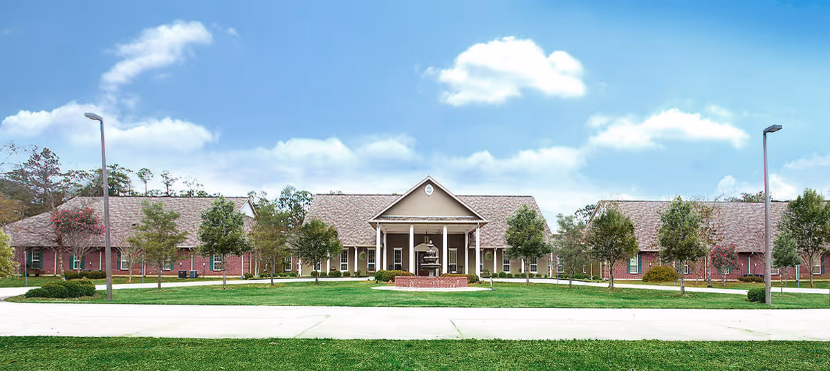 Front exterior of a single-story brick senior living building with a central portico, lawn, trees, and lamp posts under a blue sky.