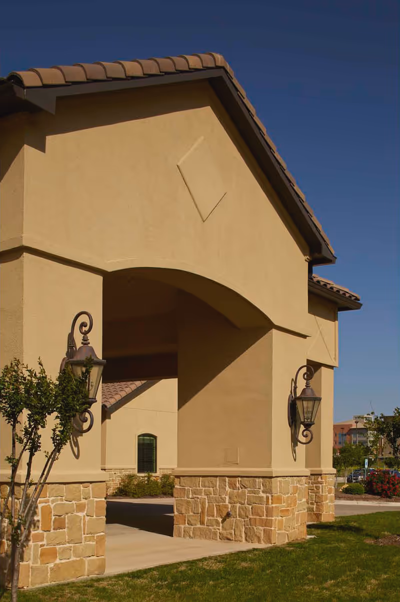 Exterior view of a tan building entrance with an arched portico and stone base.
