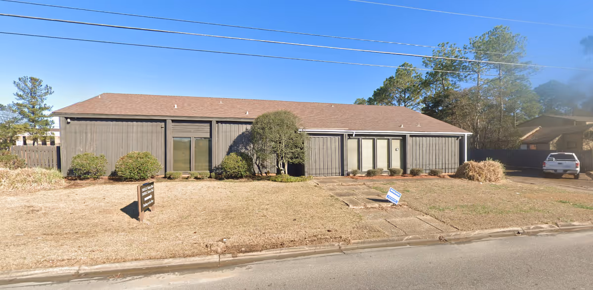 Single story gray building with a brown roof, shrubs, and a driveway in a suburban lot.
