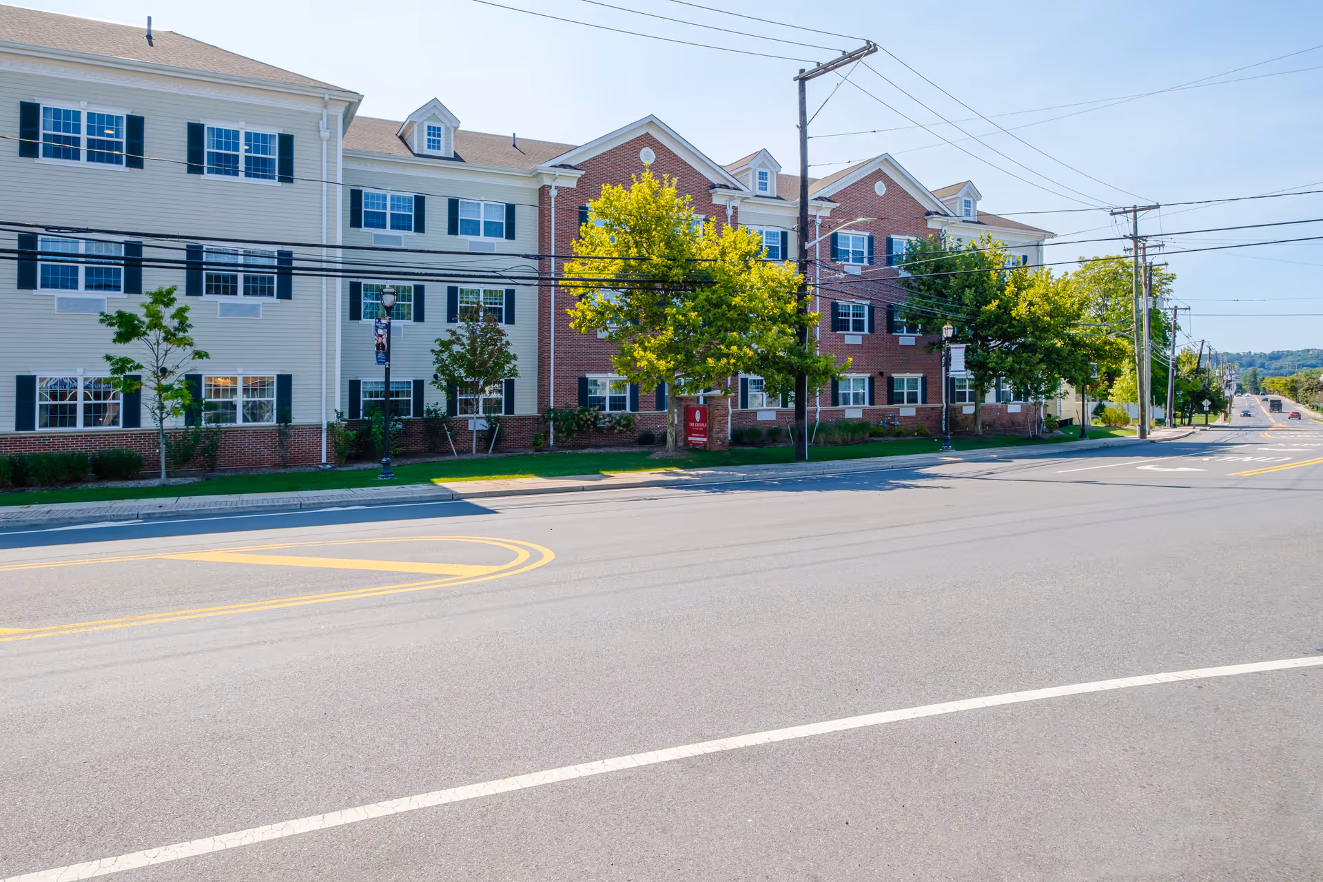 Front exterior of a multi-story senior living building with brick and siding facade, trees, and power lines along a street.