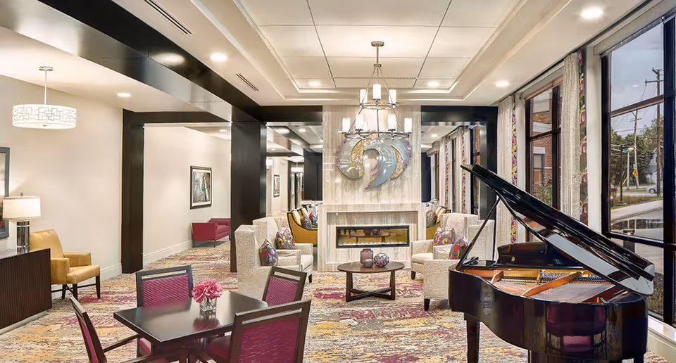 Bright communal lounge with seating, a fireplace, a grand piano and dining tables near large windows.