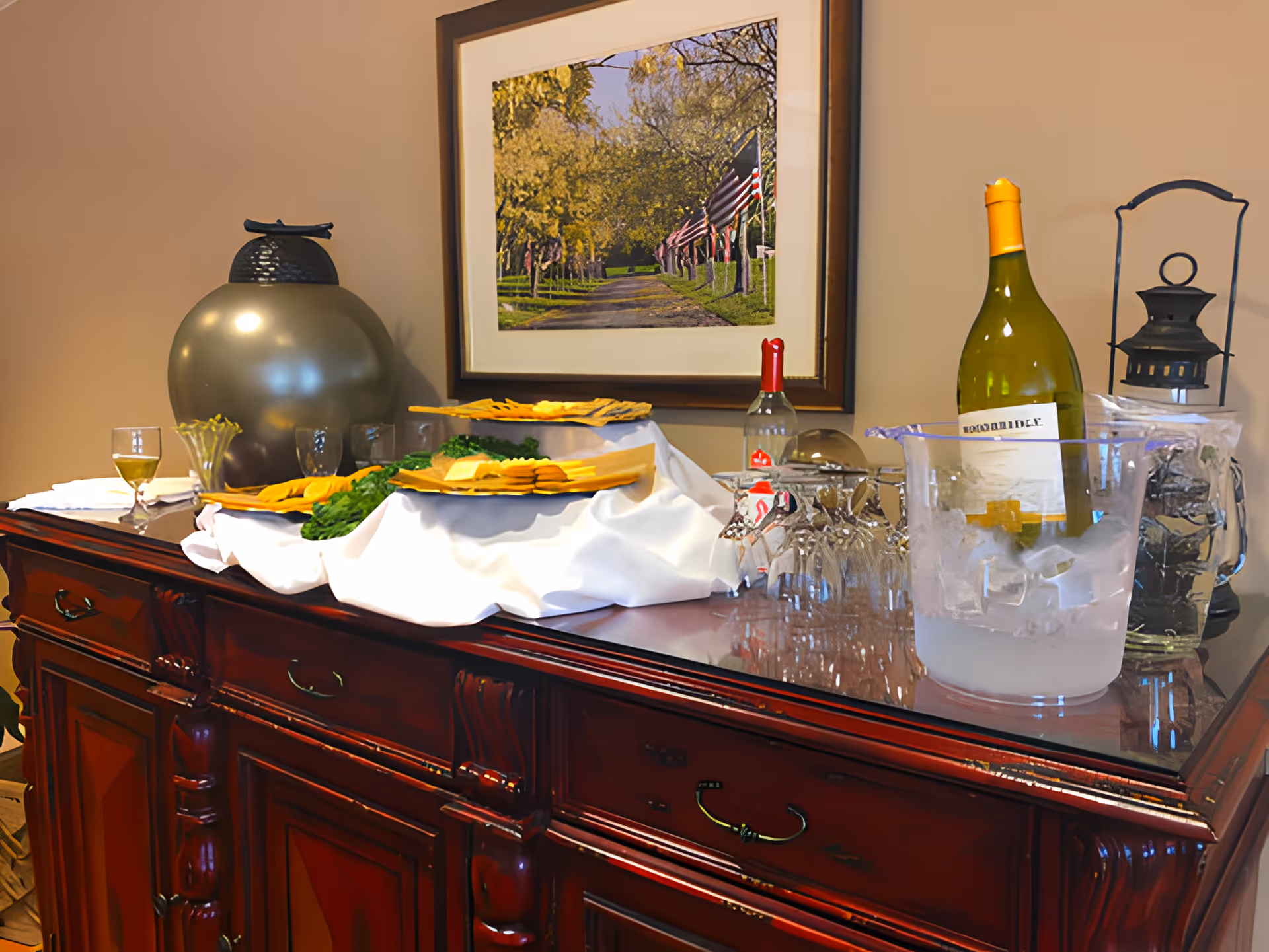 A polished wooden sideboard topped with wine bottles, an ice bucket, glassware, plates of appetizers and decorative items beneath a framed photograph.