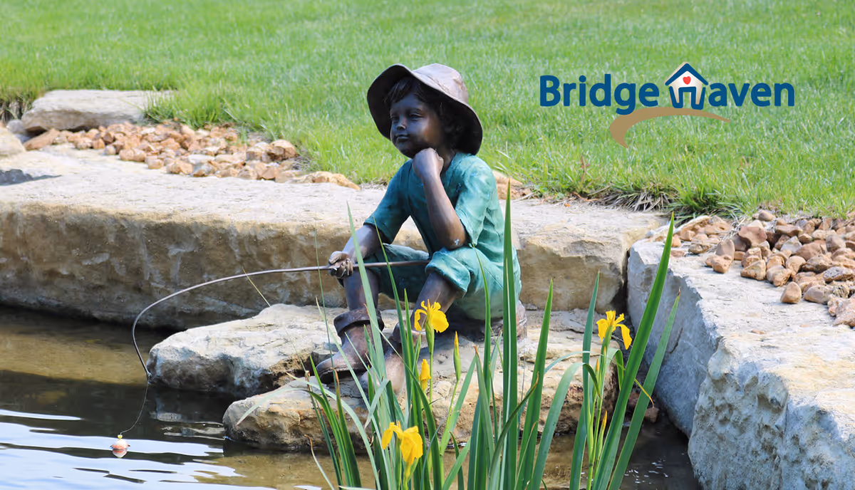 Statue of a child sits by a pond with rocks, flowers, and Bridge Haven logo.