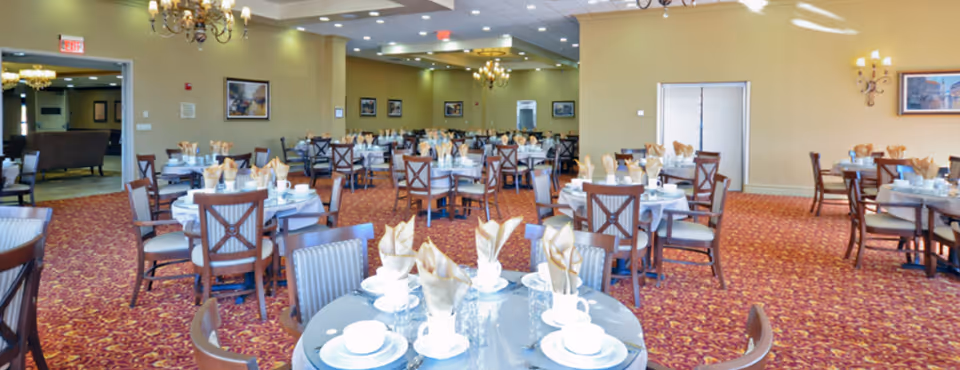 A spacious dining room with multiple round tables set with white plates, cups, glasses, and beige folded napkins. The room has a red patterned carpet, beige walls adorned with framed artwork, and chandeliers hanging from the ceiling. There is an open doorway leading to another room with additional seating.