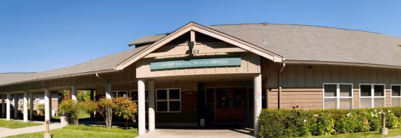 Single-story building with a covered entrance, green sign, and manicured shrubs under a blue sky.