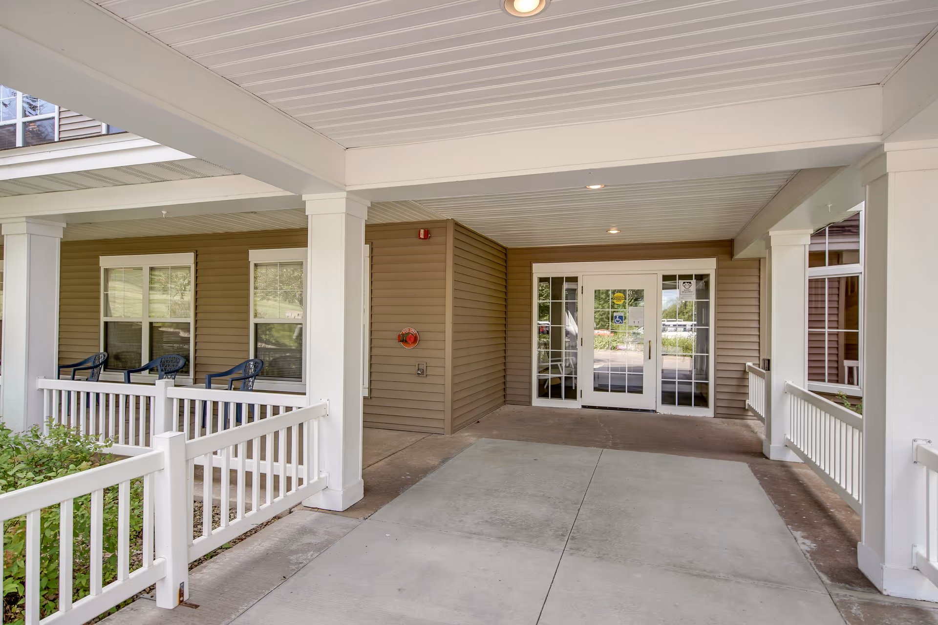 Covered entrance area of Oak Ridge Assisted Living with white pillars and railings, beige siding walls, glass double doors with accessibility signs, and three blue chairs placed near windows on the left side.