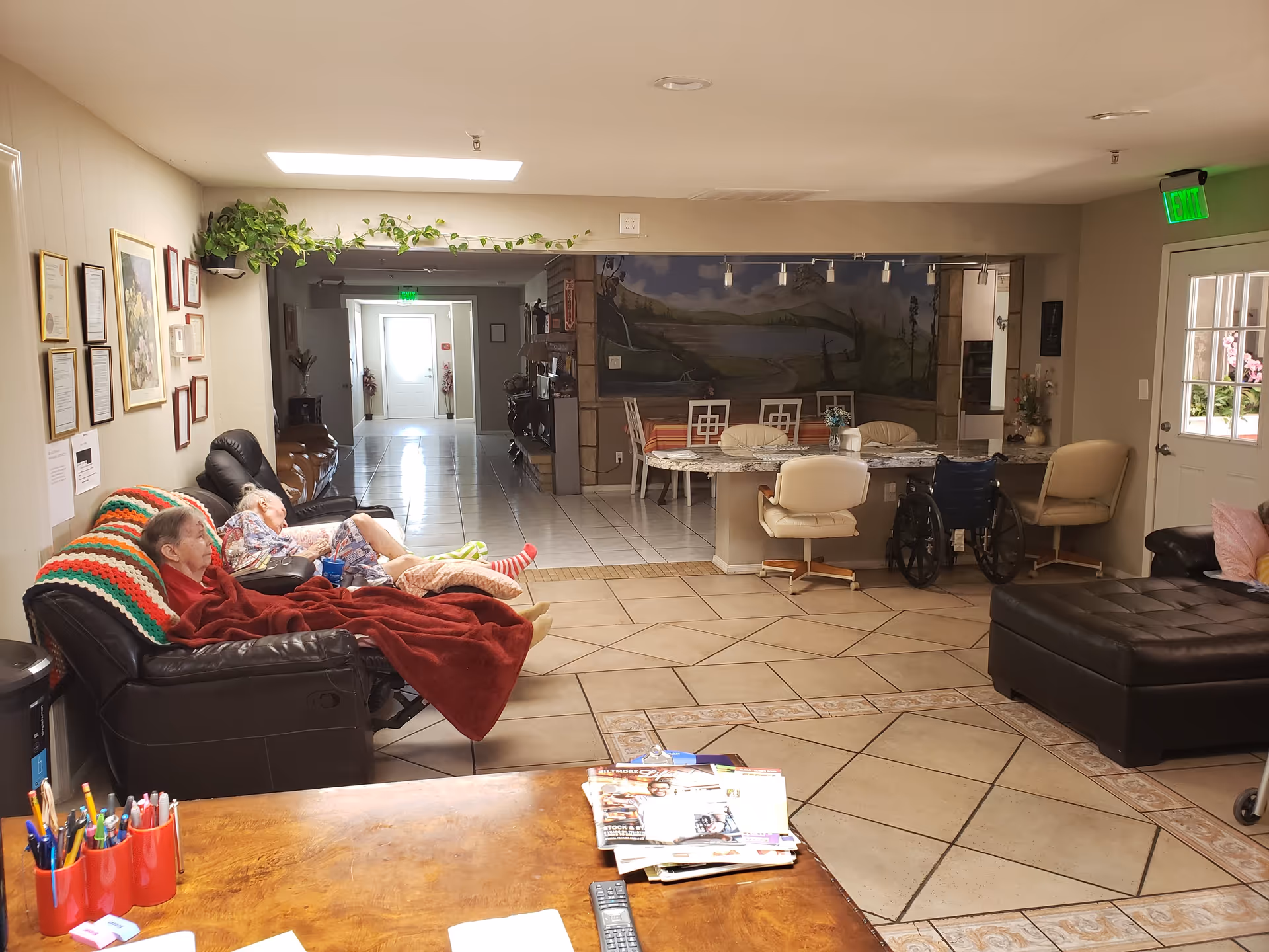 Communal living area in a senior care home with two elderly residents reclining on sofas, a dining table and chairs, a wheelchair, and a hallway leading to an exit.
