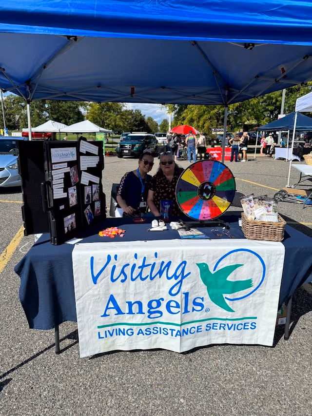 Two smiling women run a Visiting Angels booth under a blue canopy at an outdoor event.