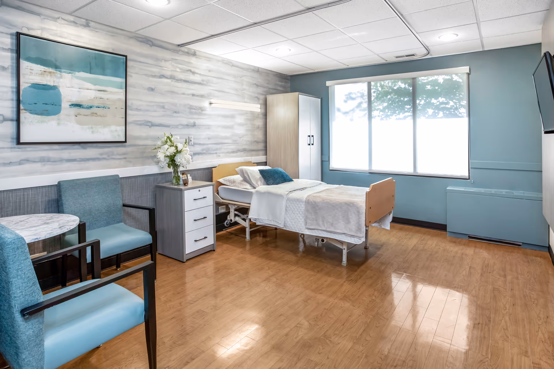 Bright patient bedroom in a senior facility with bed, seating, and large window.