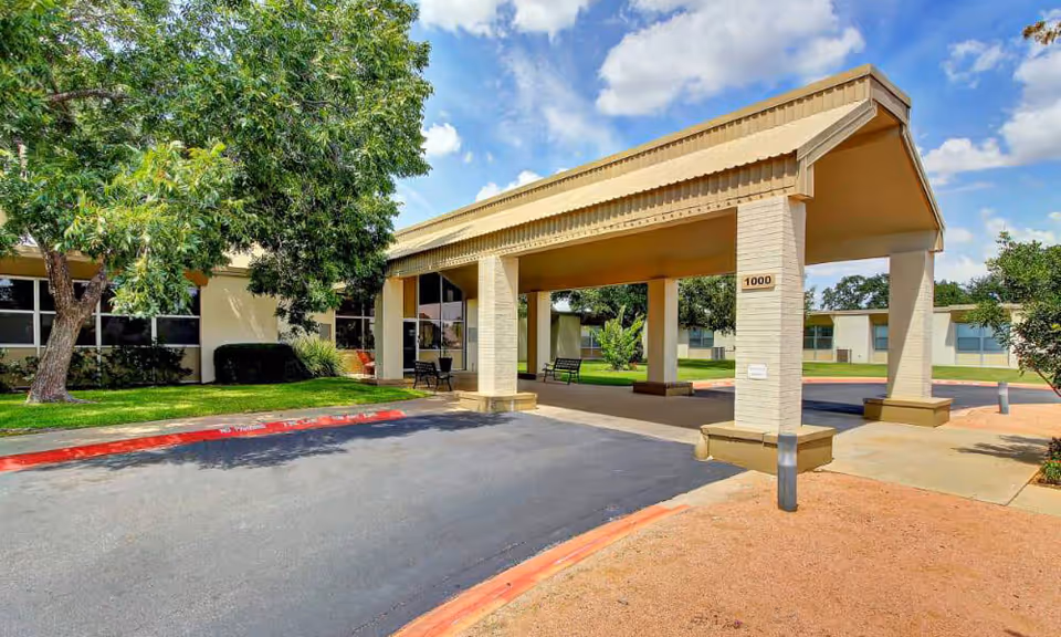 Covered entry porte-cochère and driveway of a single-story senior care building with benches and trees.