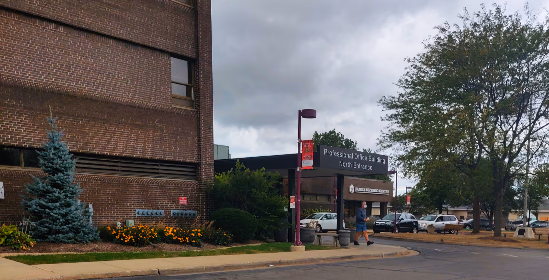 Brick exterior of a medical facility with entrance sign, cars parked nearby, and pedestrians.