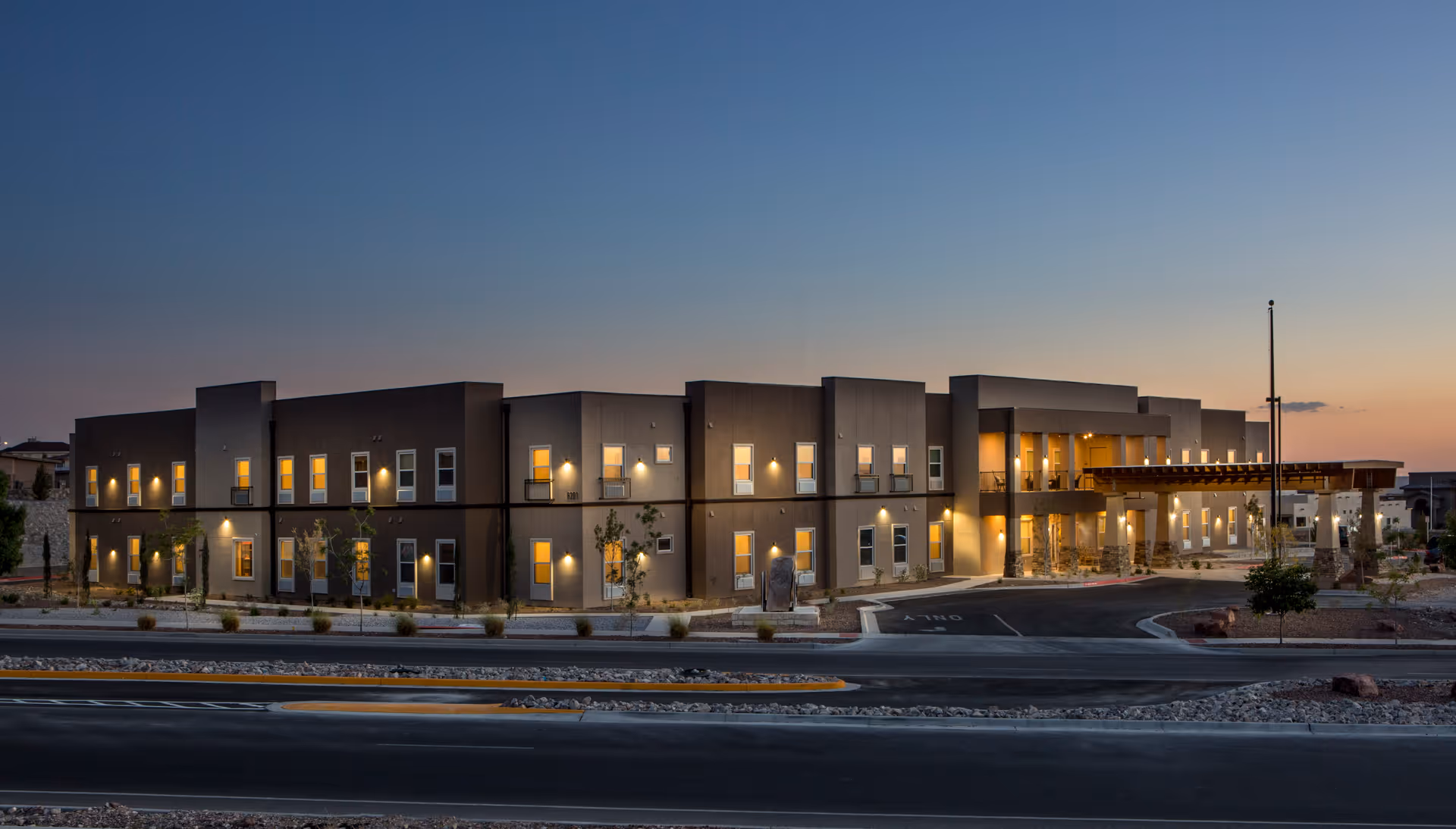 Exterior view of The Legacy at Cimarron senior living facility at dusk, showing a modern two-story building with multiple windows illuminated from inside, a covered entrance with lights, and a driveway in front.