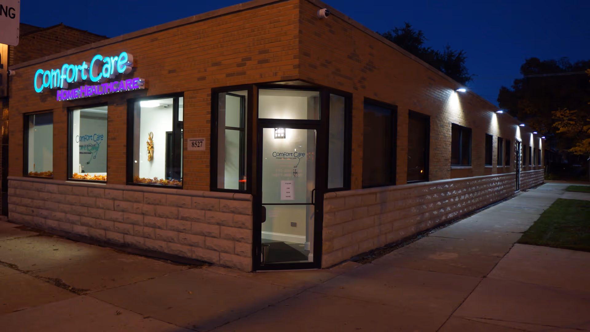 Night exterior of Comfort Care Home Healthcare facility with illuminated sign and glass entrance.