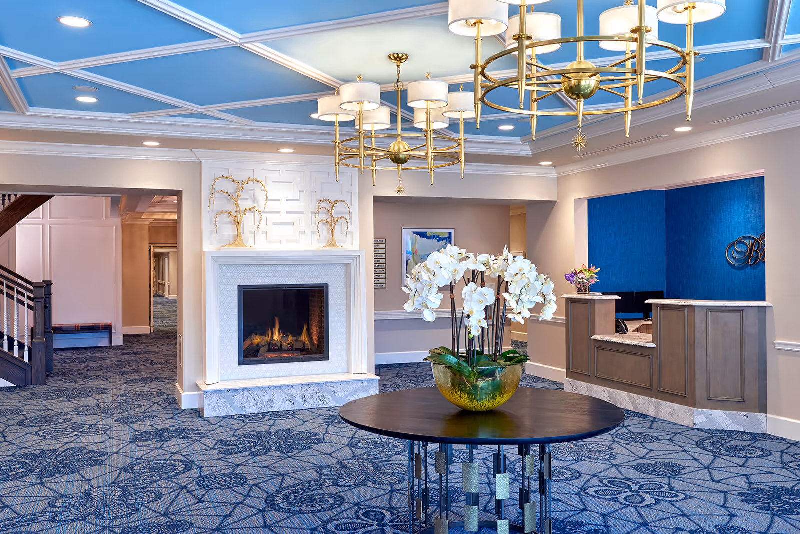 A spacious and elegant assisted living facility lobby with a blue patterned carpet, a round table with a large vase of white orchids in the center, a fireplace with decorative golden tree sculptures above it, and a reception desk with a blue accent wall behind it. The ceiling features blue panels with white trim and gold chandeliers.