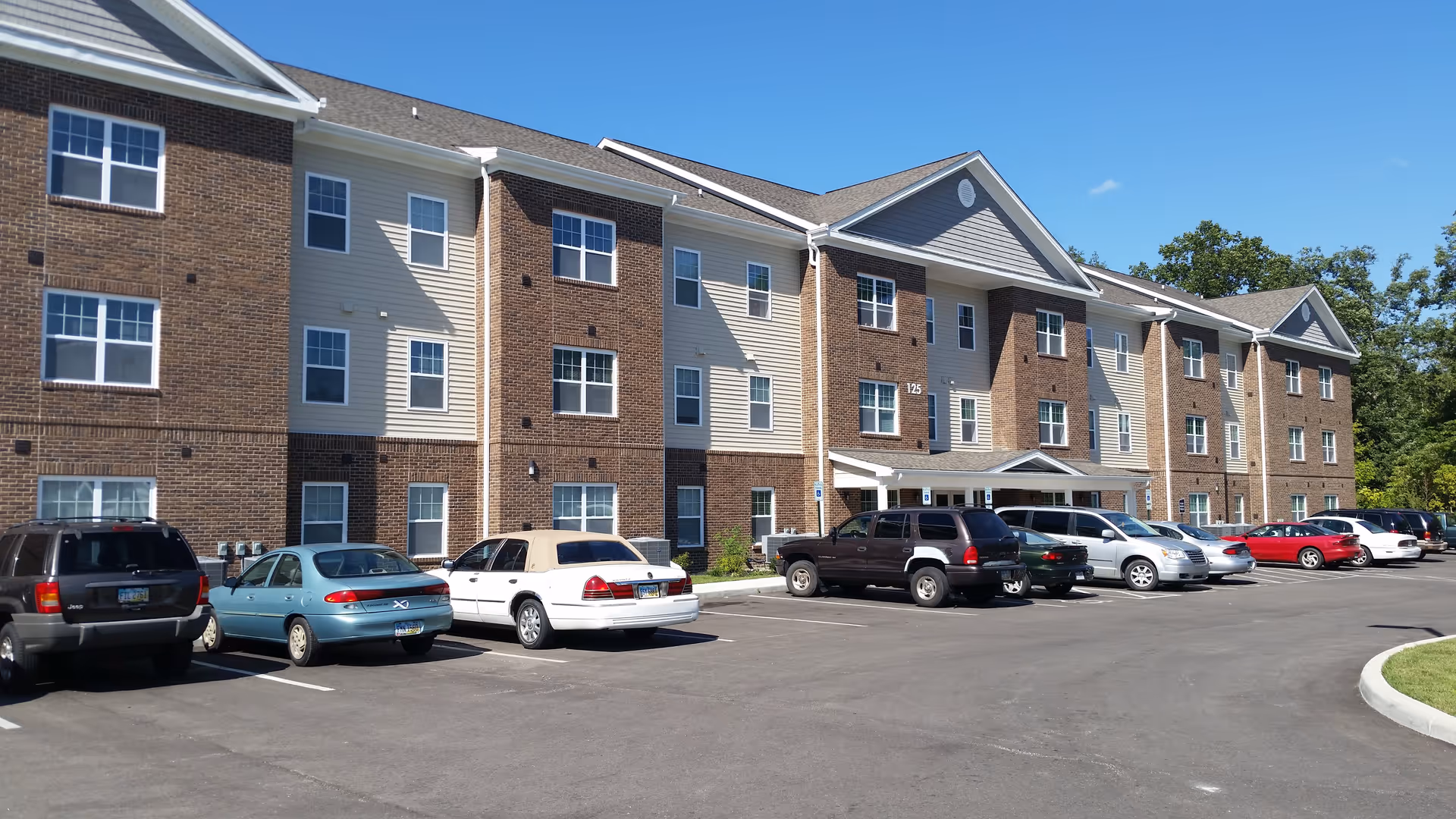 Exterior view of a three-story residential building with brick and siding facade under a clear blue sky. Several parked cars are visible in front of the building, which has multiple windows and a covered entrance.