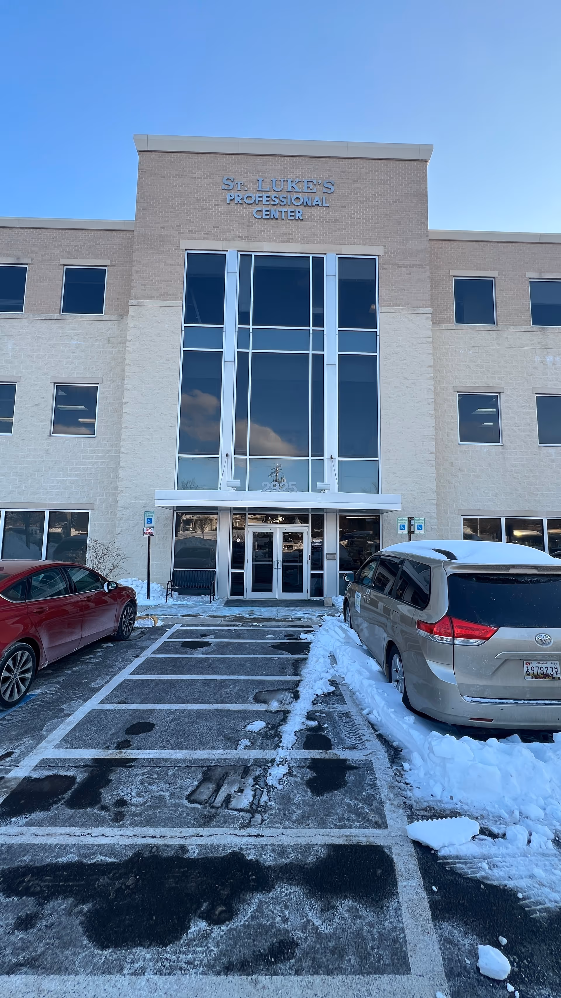 Front of a beige-brick building with glass entry doors and parked cars in snow.