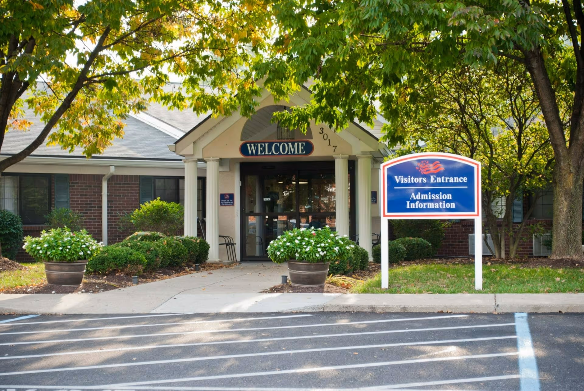 Entrance of a building with a covered porch supported by white columns, a 'WELCOME' sign above the door, and a blue sign nearby that reads 'Visitors Entrance Admission Information'. The area is surrounded by green trees and bushes with a parking lot in the foreground.