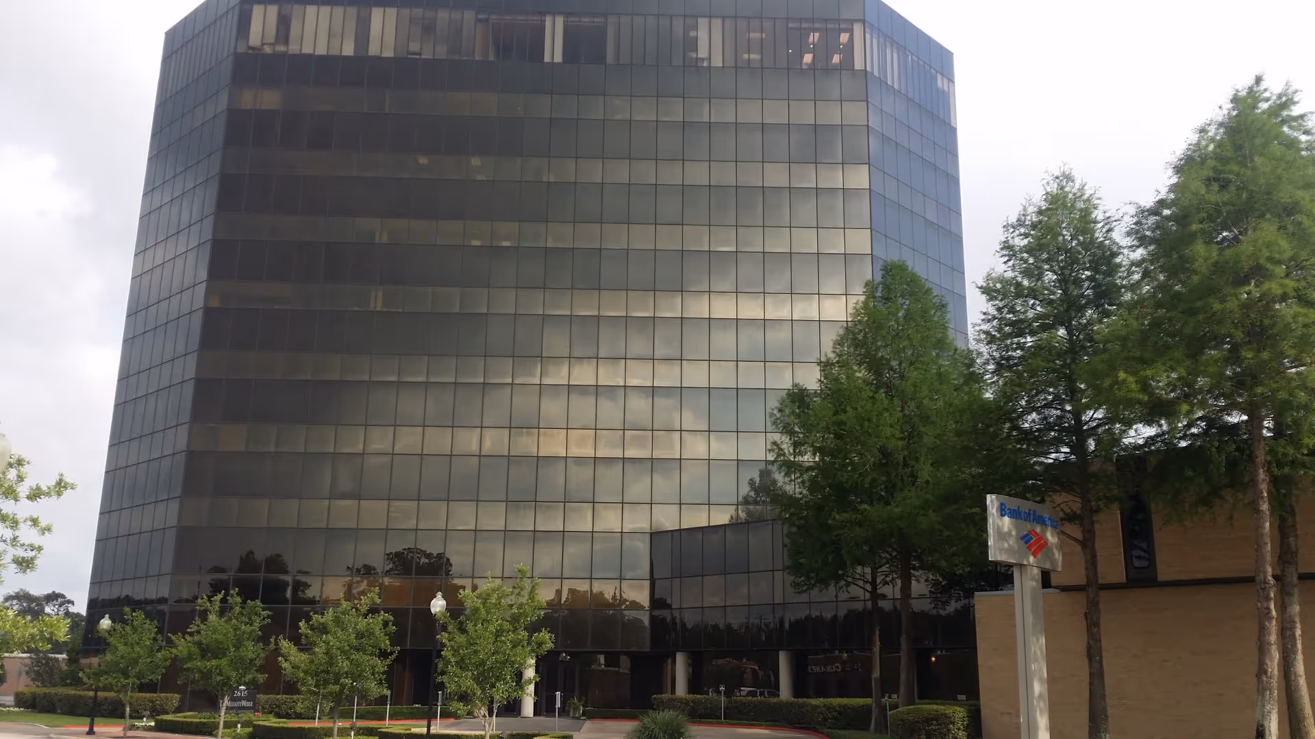 Tall glass office building with trees in foreground and a bank sign nearby.