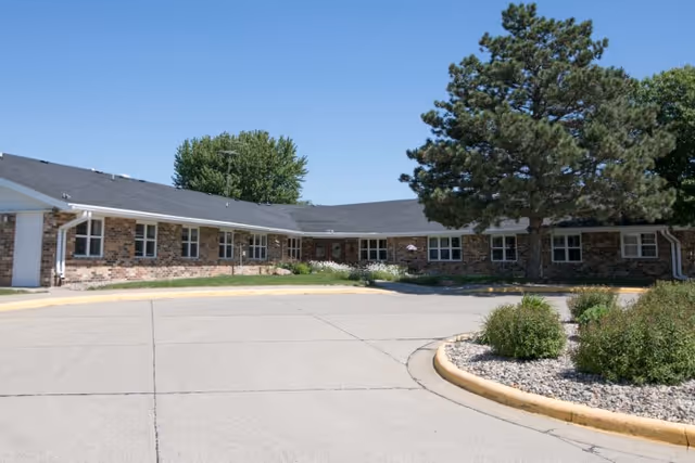 Single-story brick building with multiple windows, surrounded by trees and shrubs, under a clear blue sky. There is a curved driveway and landscaped area with bushes and rocks in front.