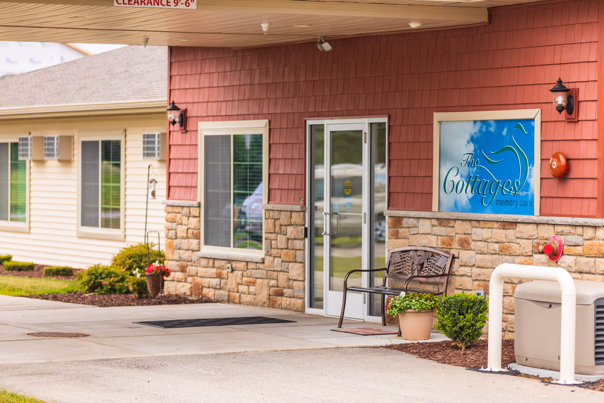 Covered entrance to The Cottages memory care showing glass double doors, a bench, potted plants, and stone-and-red siding.