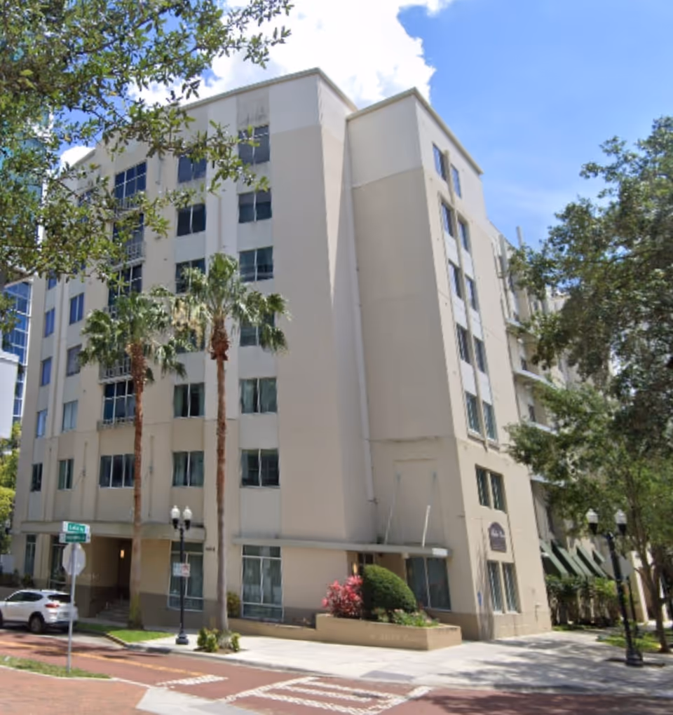 Tall, light-colored senior living building facade with palm trees and a street in front.