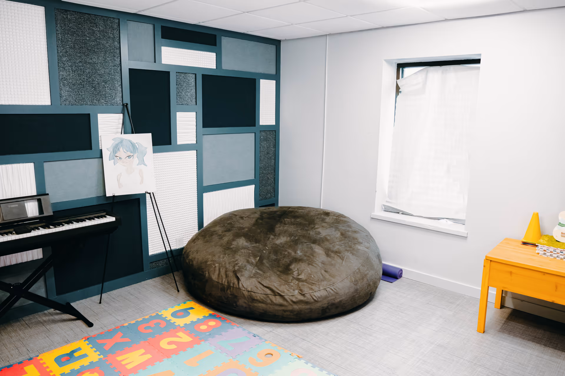 Interior activity room with acoustic panels, keyboard, large bean bag, and colorful play mat.