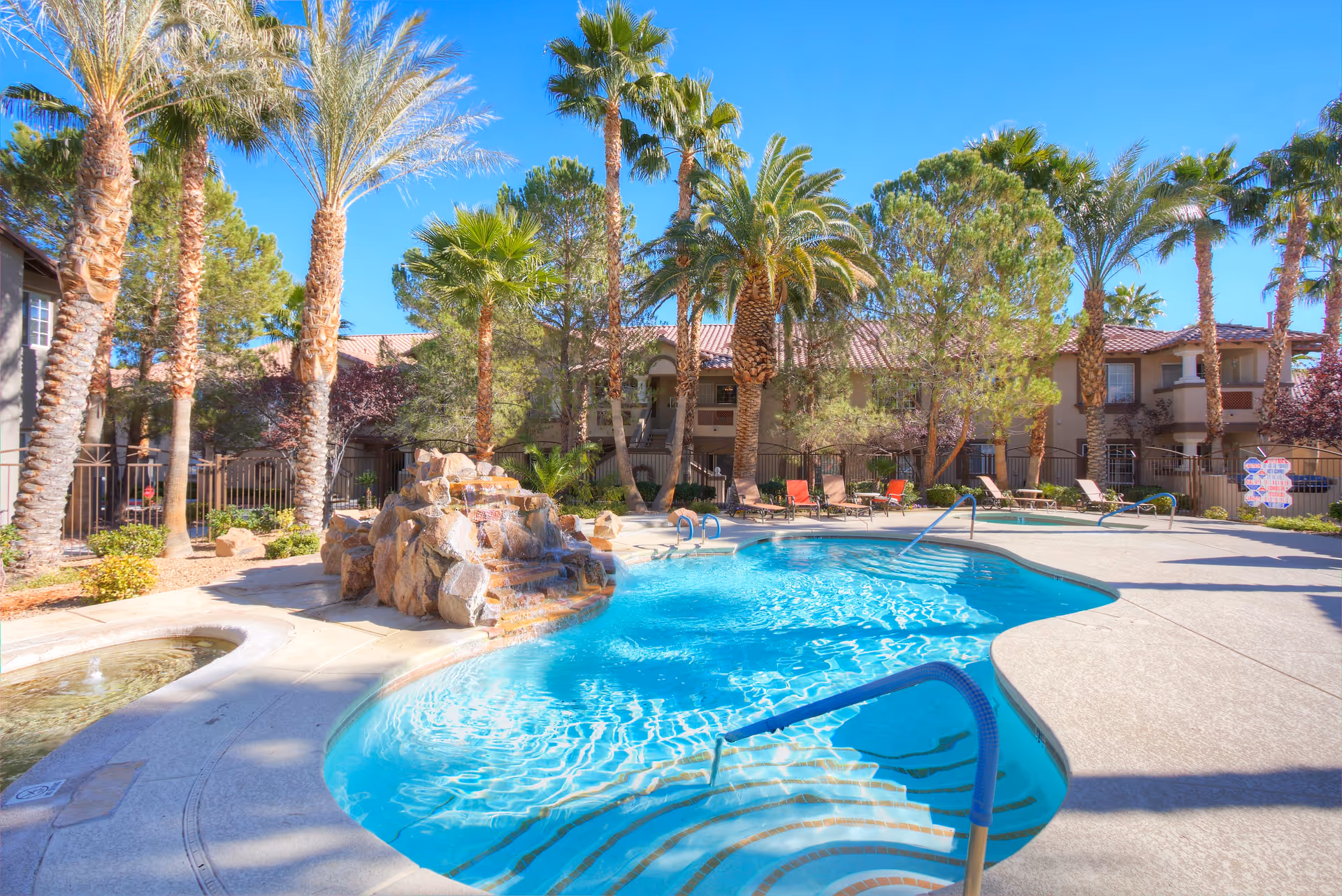 Outdoor swimming pool area with clear blue water, surrounded by palm trees and lounge chairs. There is a rock waterfall feature flowing into the pool. In the background, there are two-story residential buildings with balconies under a clear blue sky.