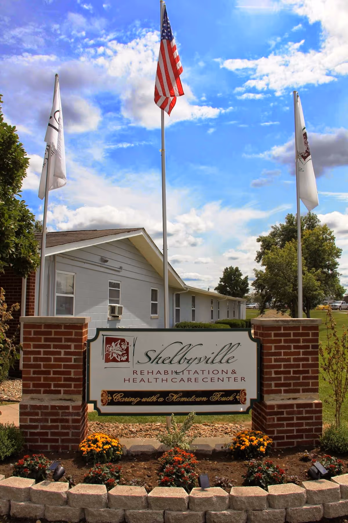 Outdoor view of Shelbyville Rehabilitation & Health Care Center sign with three flagpoles behind it, including an American flag. The building is visible in the background under a partly cloudy sky, surrounded by greenery and flowers.