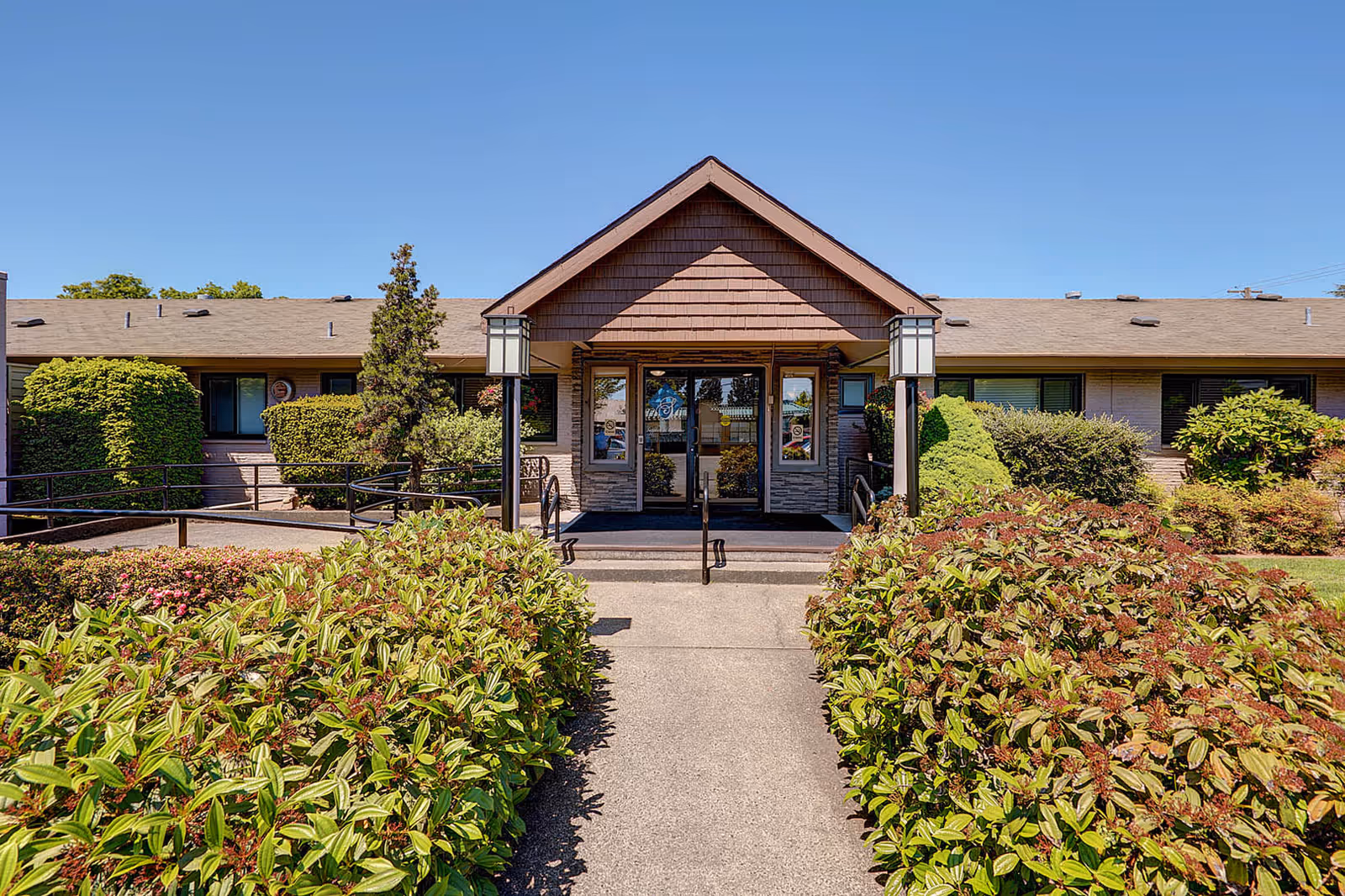 Front entrance of a single-story senior living building with a peaked entryway, walkway and landscaped shrubs.