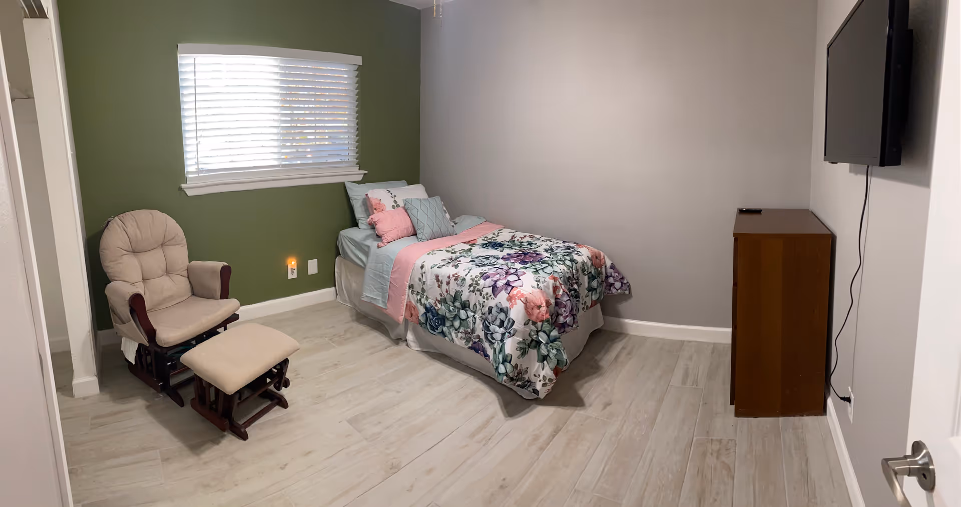 A cozy bedroom with a floral quilt, green wall, chair, and wall-mounted TV.