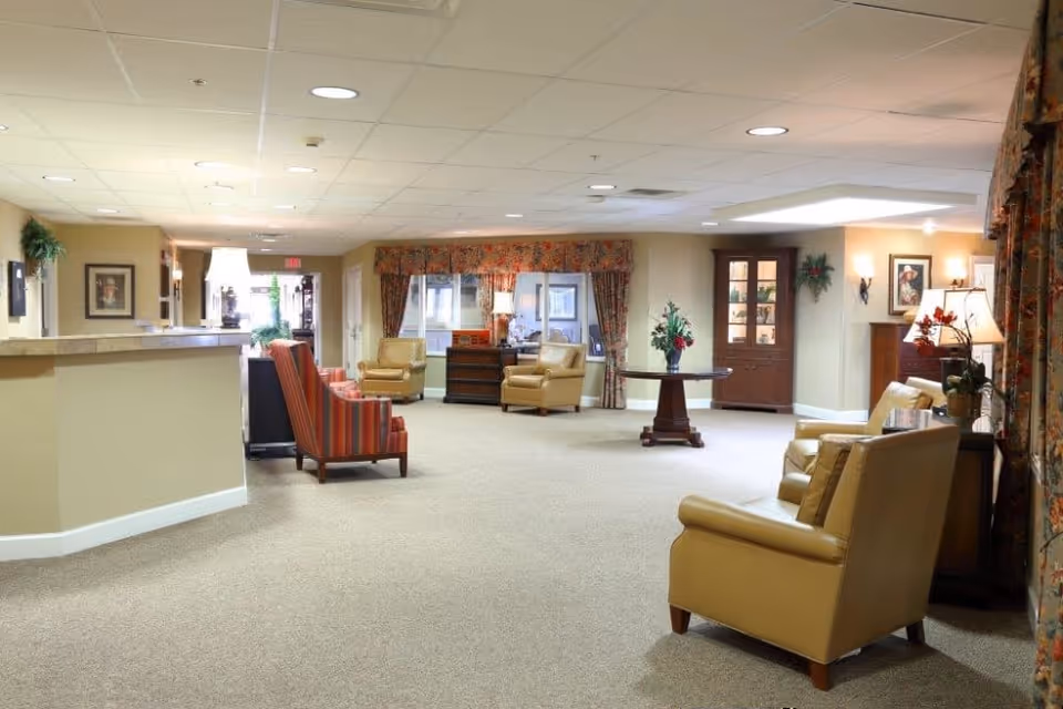 A spacious, well-lit common area with beige carpet and walls, featuring several comfortable armchairs in tan and striped upholstery. There is a round wooden table with a flower arrangement in the center, a wooden cabinet with glass doors, framed artwork on the walls, and large windows with floral curtains allowing natural light to enter.