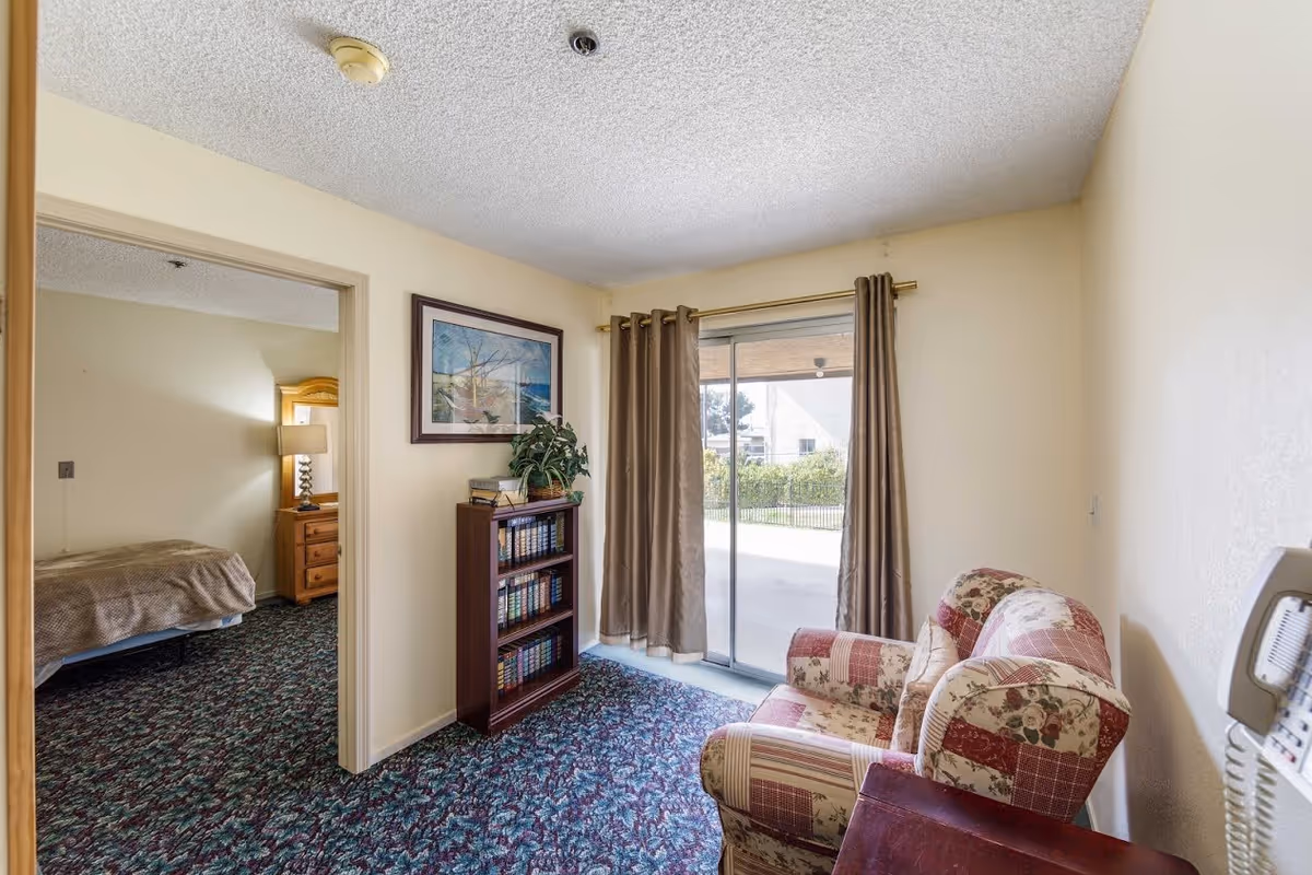 A small living area with a floral patterned armchair, a wooden bookshelf filled with books, and a framed painting on the wall. The room has a sliding glass door with brown curtains leading to an outdoor patio. Through an open doorway, a bedroom with a bed, nightstand, and lamp is visible.
