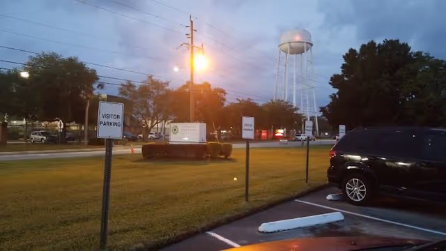 Parking lot area at dusk with signs indicating visitor parking. A black SUV is parked in the lot. In the background, there is a water tower and a sign for Healthcare And Rehab Of Sanford surrounded by trees and grass.
