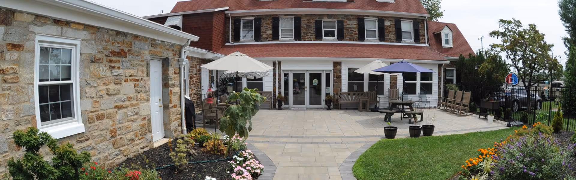 Exterior courtyard of an assisted living building with patio seating, umbrellas, potted plants, and landscaped flower beds.