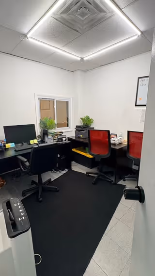 Small office workspace with two desks, computers, red chairs, plants, and a window.