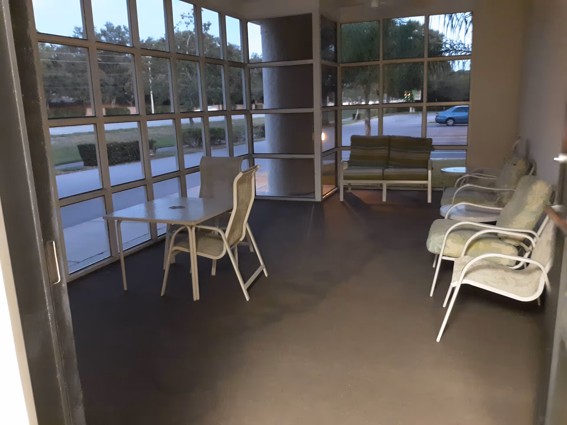 Indoor glass-enclosed lounge with white chairs, a small table, and a bench against the back wall.